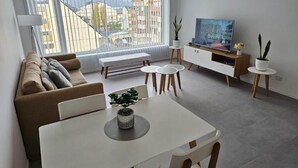 Una sala de estar moderna con un sofá, una mesa de comedor, un televisor y vistas a edificios a través de amplios ventanales.