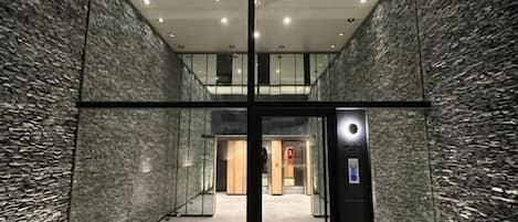An interior corridor with a glass door leading to a lobby area, featuring a textured wall and a ceiling with recessed lighting.
