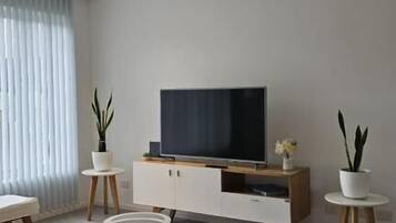 Een moderne woonkamer met een flatscreen televisie, een witte kast, een eettafel met stoelen en potplanten.