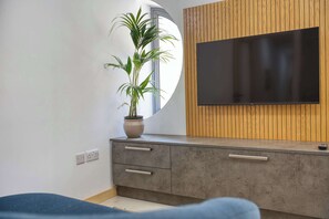Ein modernes Wohnzimmer mit einem an einer Holzpaneelwand montierten Flachbildfernseher, einer Topfpflanze auf einem grauen Schrank und einem Fenster mit Ausblick.