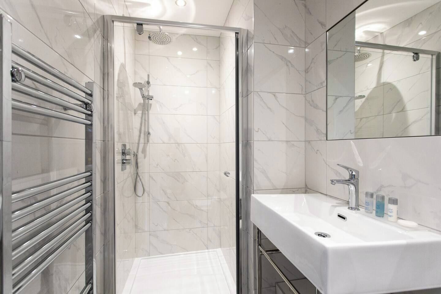A modern bathroom with a glass-enclosed shower, a white sink with a mirror, and a marble-tiled floor.