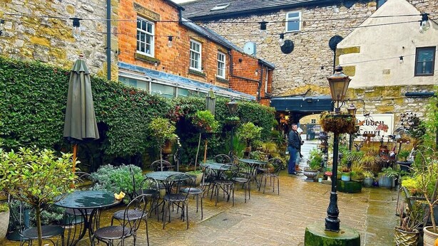 An outdoor patio area with tables and chairs, surrounded by potted plants and a stone floor.