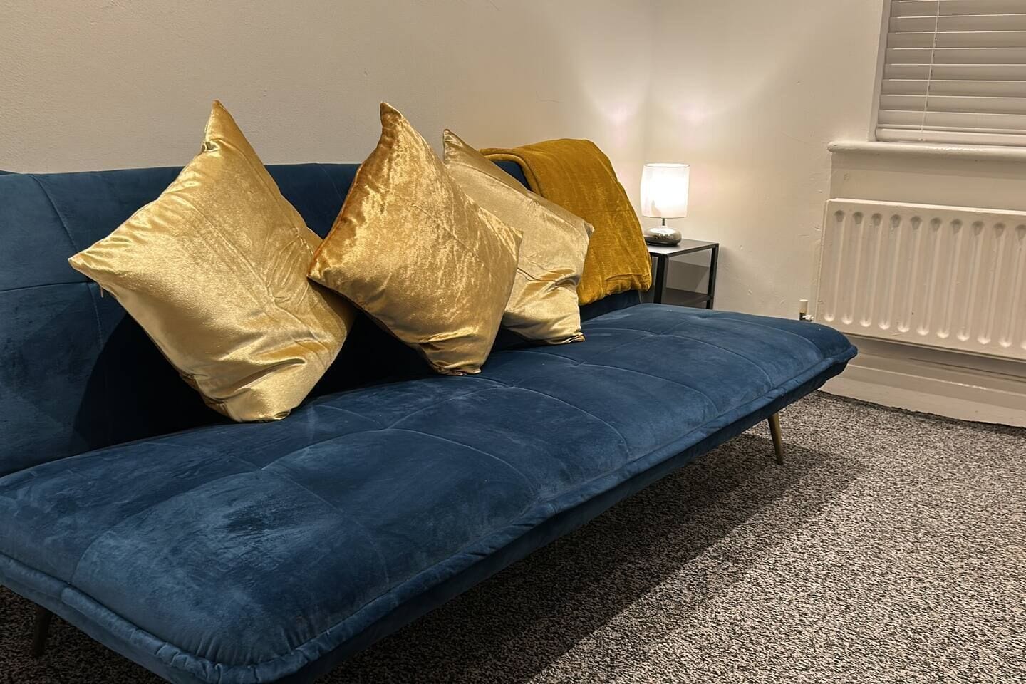 A blue sofa with four cushions, a small table with a lamp, and a radiator in a room.