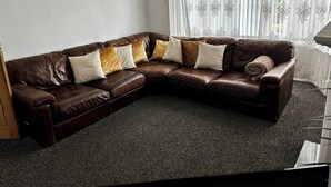 A brown leather sectional sofa with multiple cushions, a dark carpeted floor, and a window with sheer curtains.