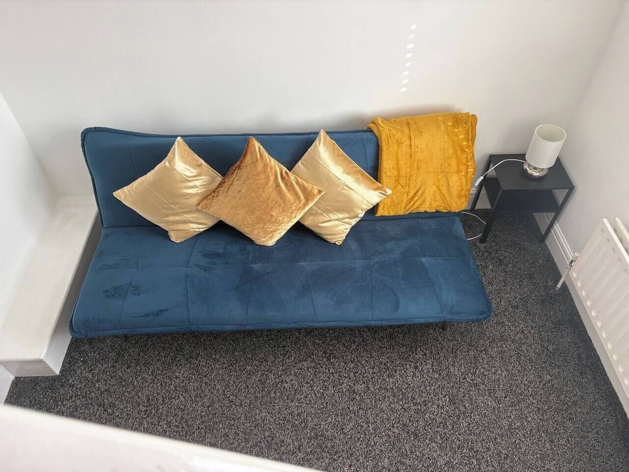 Un canapé bleu agrémenté de trois coussins dorés et d’un plaid couleur moutarde, une petite table d’appoint noire surmontée d’une lampe blanche, et un radiateur fixé au mur.
