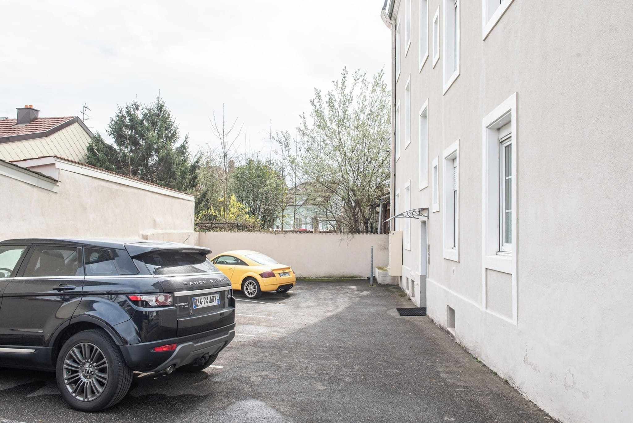 Ein Parkplatz mit zwei Autos, ein Gebäude mit weißen Wänden und Fenstern sowie Bäume im Hintergrund.
