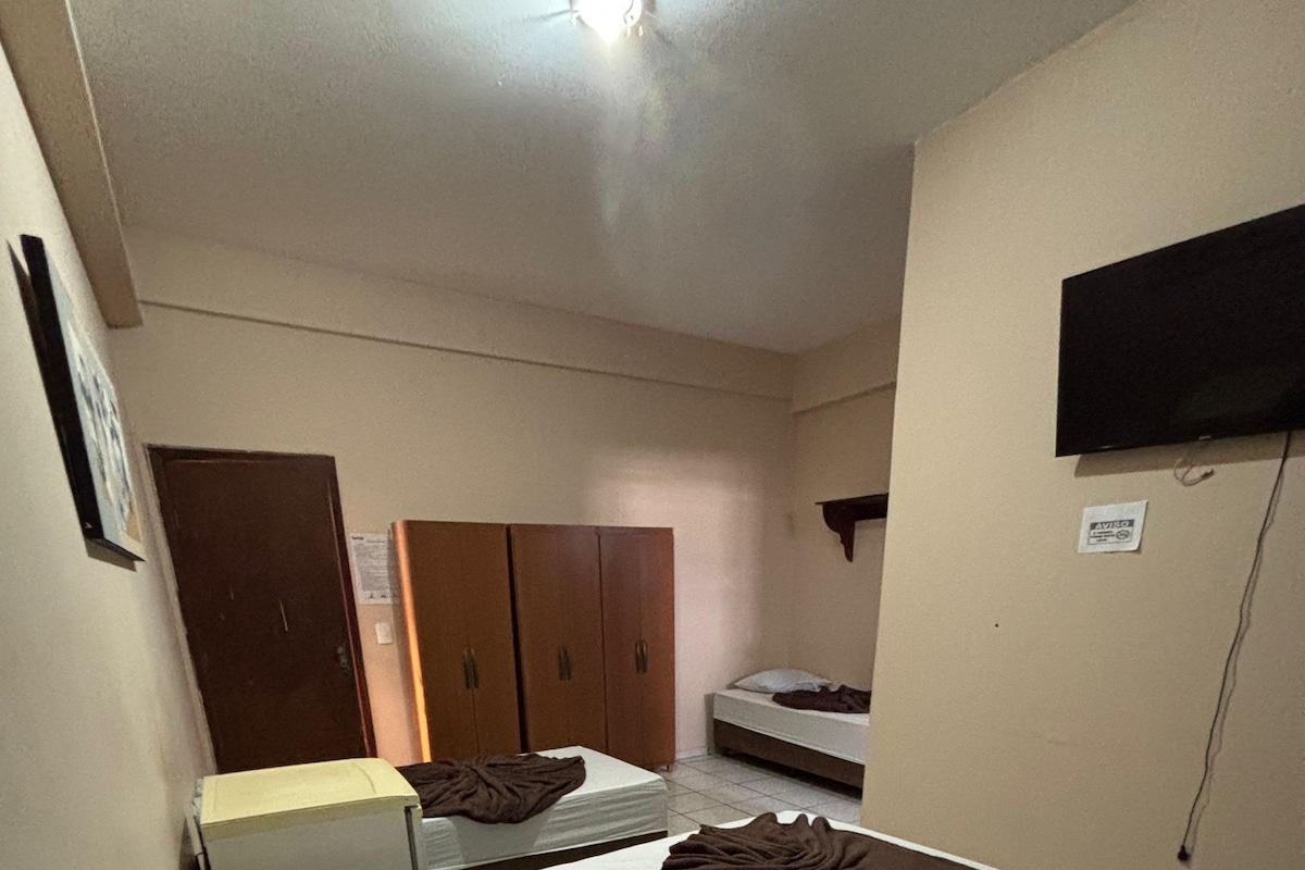Una cama individual con almohadas y una colcha, un armario, una mesita y un televisor montado en la pared.