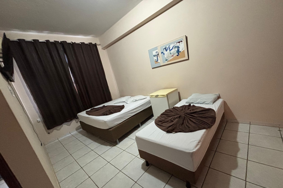 Una habitación de hotel con dos camas individuales, una mesita de noche pequeña, cortinas en una ventana y una obra de arte colgada en la pared.