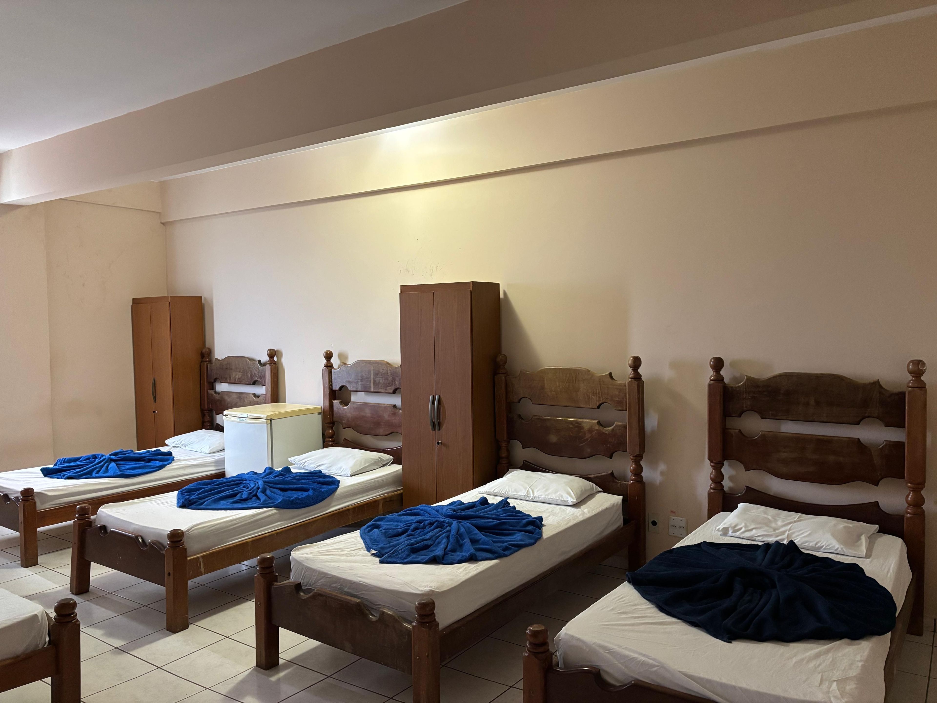 Ein Zimmer mit vier Holzbetten, jedes mit einer blauen Decke und weißen Kissen. Holzschränke und eine kleine weiße Kommode sind ebenfalls vorhanden.