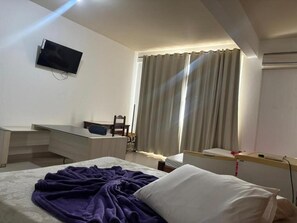 A hotel room with a bed, a desk, a chair, a TV mounted on the wall, and a window with curtains. - HOTEL VIA MONTES (Montes Claros)