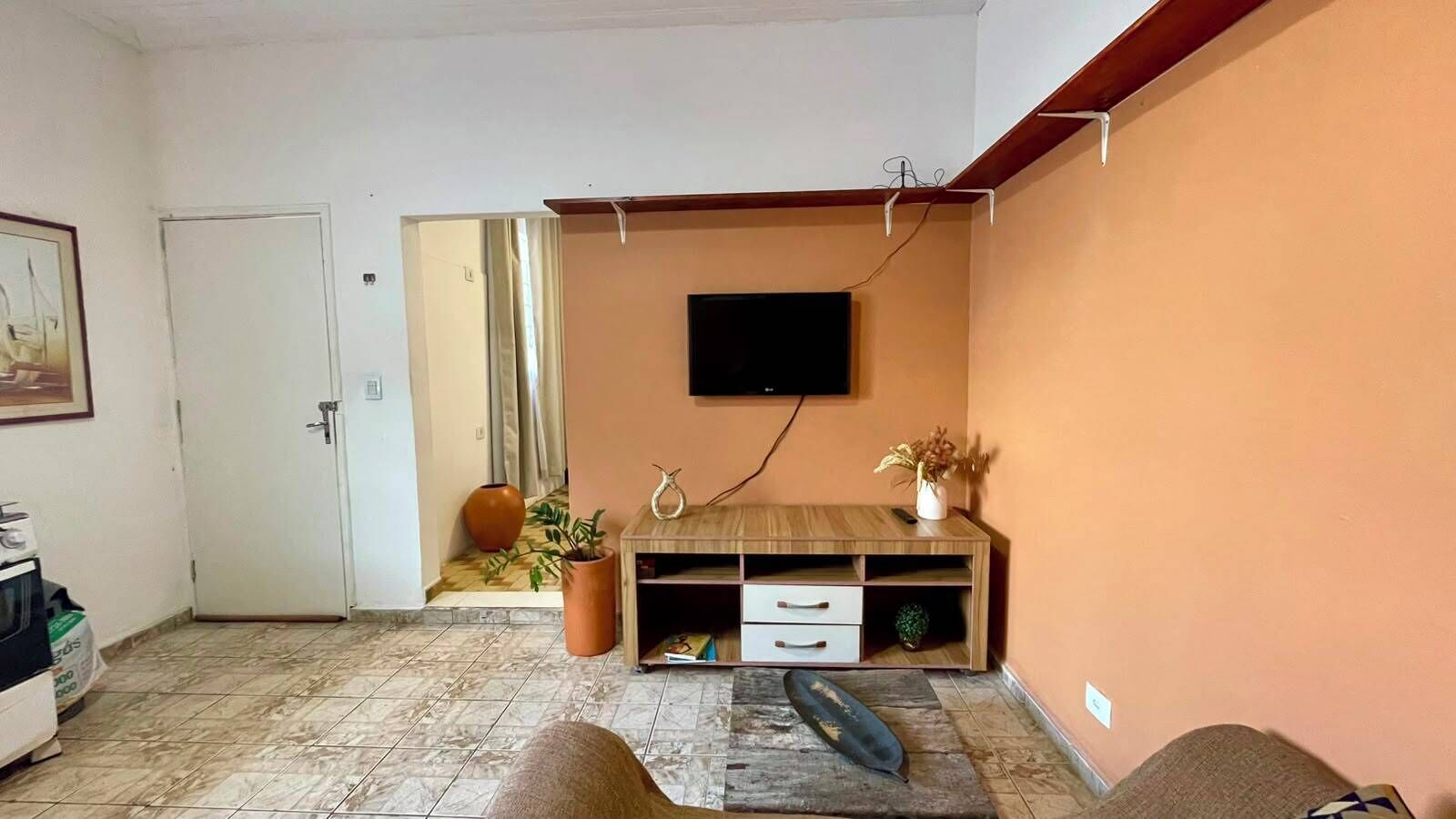 Uma sala de estar com televisão de tela plana na parede, um móvel de madeira com gavetas, um vaso de planta e uma porta que dá para outro cômodo.
