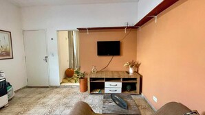 Uma sala de estar com televisão de tela plana na parede, um móvel de madeira com gavetas, um vaso de planta e uma porta que dá para outro cômodo.
