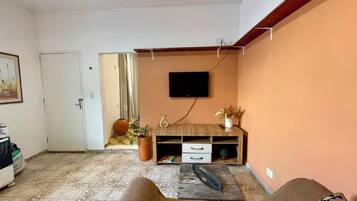 Uma sala de estar com televisão de tela plana na parede, um móvel de madeira com gavetas, um vaso de planta e uma porta que dá para outro cômodo.