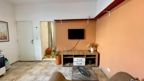 Ein Wohnzimmer mit einem an der Wand befestigten Flachbildfernseher, einer Holz-Unterhaltungsanlage mit Schubladen, einer Topfpflanze und einer Tür zu einem anderen Raum.