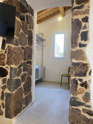 A stone-walled room with a TV mounted on the wall, a window, and a chair. - Antony's House (Orosei)