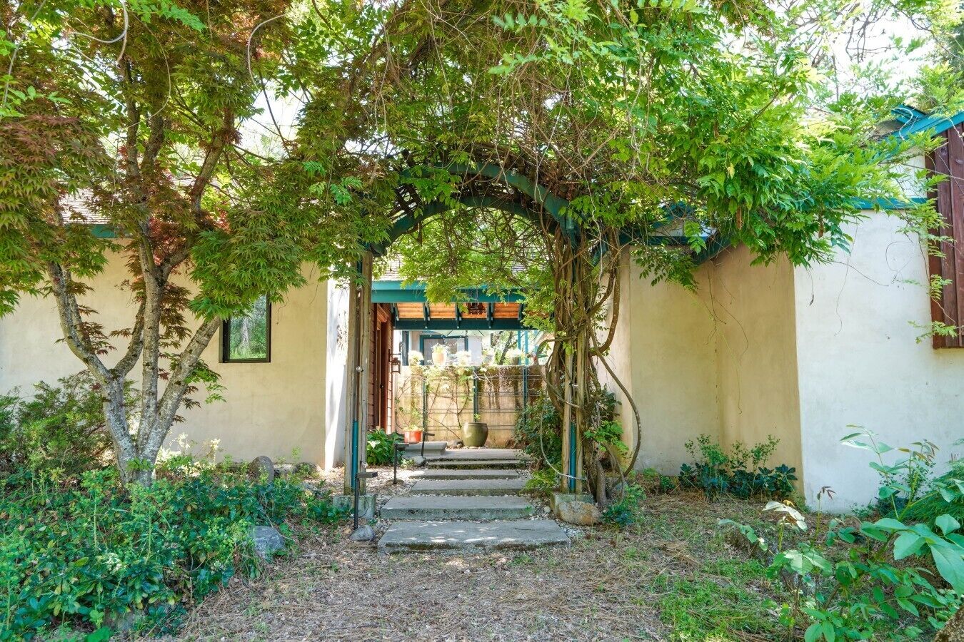 Un sentier de jardin menant à l’entrée d’un bâtiment, avec une arche en bois, le tout entouré de verdure et agrémenté d’un escalier en pierre.