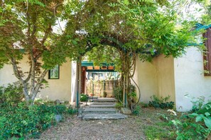 Ein Gartenweg führt zu einem Gebäudeeingang mit einem hölzernen Bogen, umgeben von Grün und einer Steintreppe.