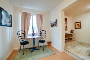 Ein Zimmer mit Tisch und zwei Stühlen, einem Fenster mit Vorhängen, einem Bücherregal und einem gerahmten Bild an der Wand.