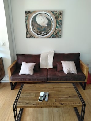 A living room with a brown leather sofa, two white cushions, a wooden coffee table with a remote control, and a large abstract painting on the wall. - L'Instant Sud in Châteauneuf-de-Grasse (Châteauneuf-Grasse)