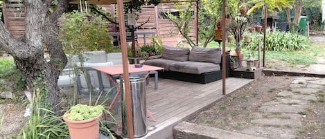 A covered outdoor seating area with a table and sofa, surrounded by potted plants and a wooden deck.