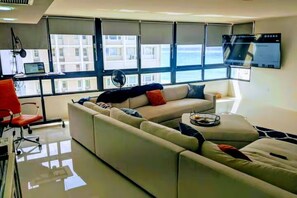 Ein modernes Wohnzimmer mit einem großen Sofa, einem roten Sessel, einem Flachbildfernseher und Blick auf Gebäude durch große Fenster.