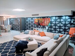 Ein modernes Wohnzimmer mit einer Eckcouch, einem Essbereich mit Stühlen und einem großen abstrakten Gemälde an der Wand.