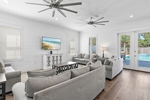 A spacious living room with grey sofas, a TV, ceiling fans, and a view of a swimming pool. - Saltwater Sanctuary | Billiards, Spa, & Golf Cart (Miramar Beach)