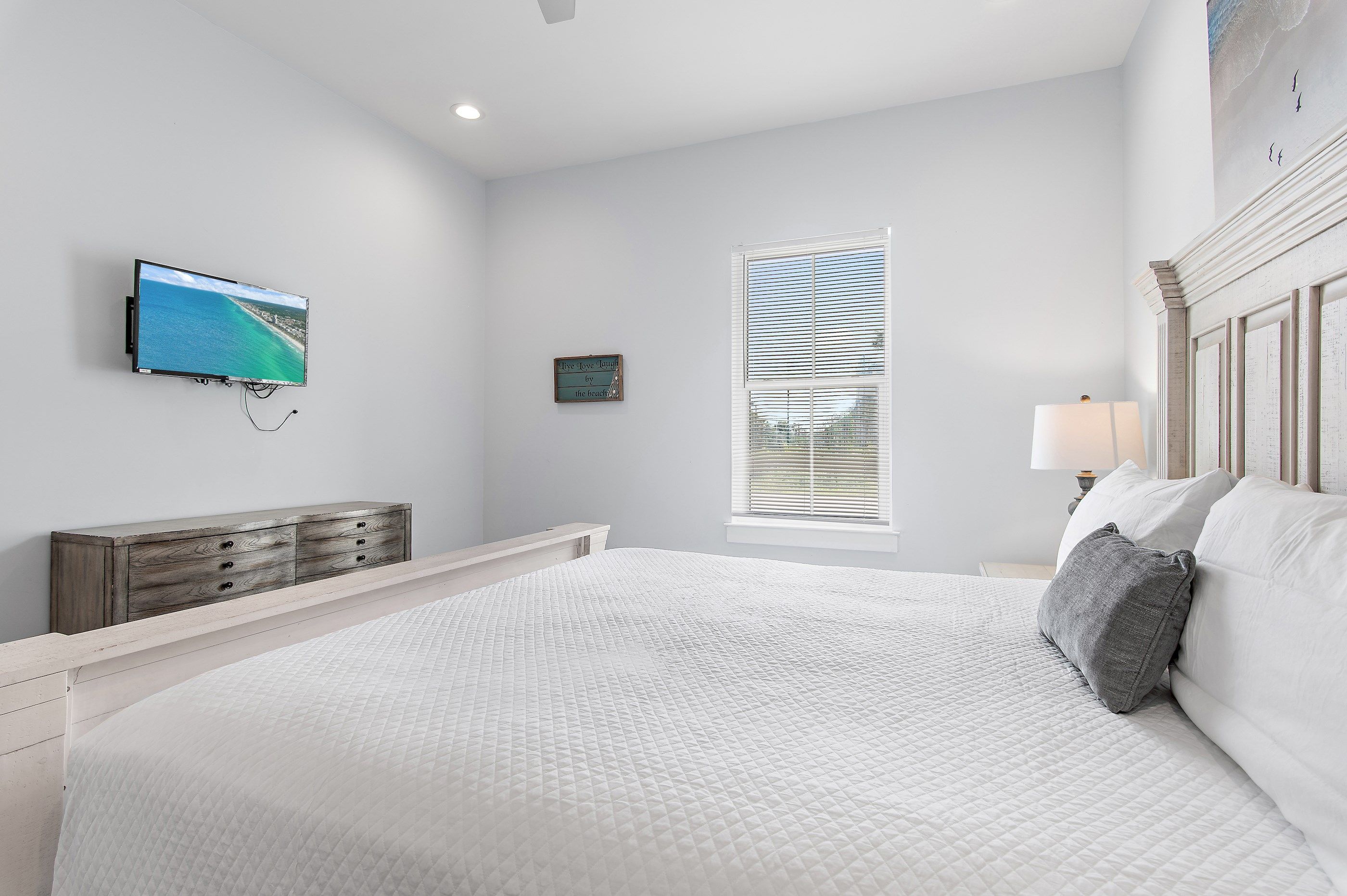 Ein Schlafzimmer mit einem großen Bett, einem an der Wand montierten Fernseher, einem Fenster mit Jalousien und einem kleinen Nachttisch mit Lampe.