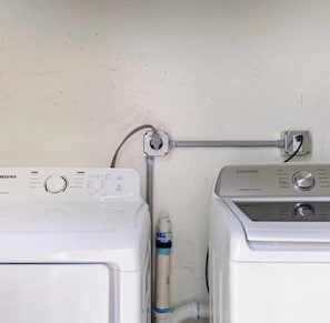 Zwei weiße Samsung Waschmaschinen stehen nebeneinander, verbunden durch einen grauen Schlauch und mit einer an der Wand montierten Steckdose.