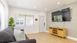 A modern living room with a flat-screen TV mounted on the wall, a wooden entertainment unit, a grey sofa, and a large window with blinds. - 3bed Near Beach, Wynwood Design District, Brickell (Miami)