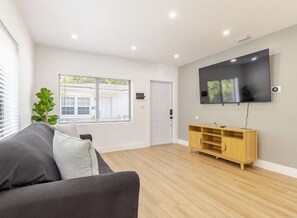 Ein modernes Wohnzimmer mit einem an der Wand befestigten Flachbildfernseher, einer Holzunterhaltungsecke, einem grauen Sofa und einem großen Fenster mit Jalousien.