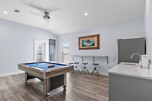 A modern room with a pool table, a ceiling fan, a bar area with stools, and a large window. - Solara Sanctuary | Golf Cart, Spa, & Billiards (Miramar Beach)