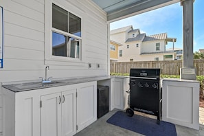A covered outdoor kitchen area with a grill, a sink, and a window overlooking a residential neighborhood. - Solara Sanctuary | Golf Cart, Spa, & Billiards (Miramar Beach)