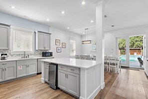 A modern kitchen with white cabinetry, a central island, and a dining area with a view of a backyard. - Solara Sanctuary | Golf Cart, Spa, & Billiards (Miramar Beach)