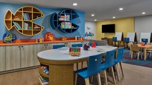 Um espaço de brincadeiras para crianças, com uma mesa redonda, cadeiras azuis e estantes cheias de brinquedos e livros.
