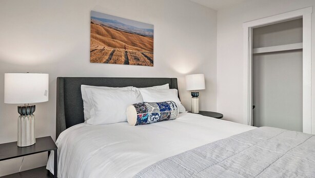 A neatly made bed with white linens and a patterned pillow, flanked by two bedside tables with lamps, in a room with a plain wall and a door. - Paso on Oak by Avantstay Cottage-style Home + Full Kitchen + Front Deck Near Downtown (Paso Robles)