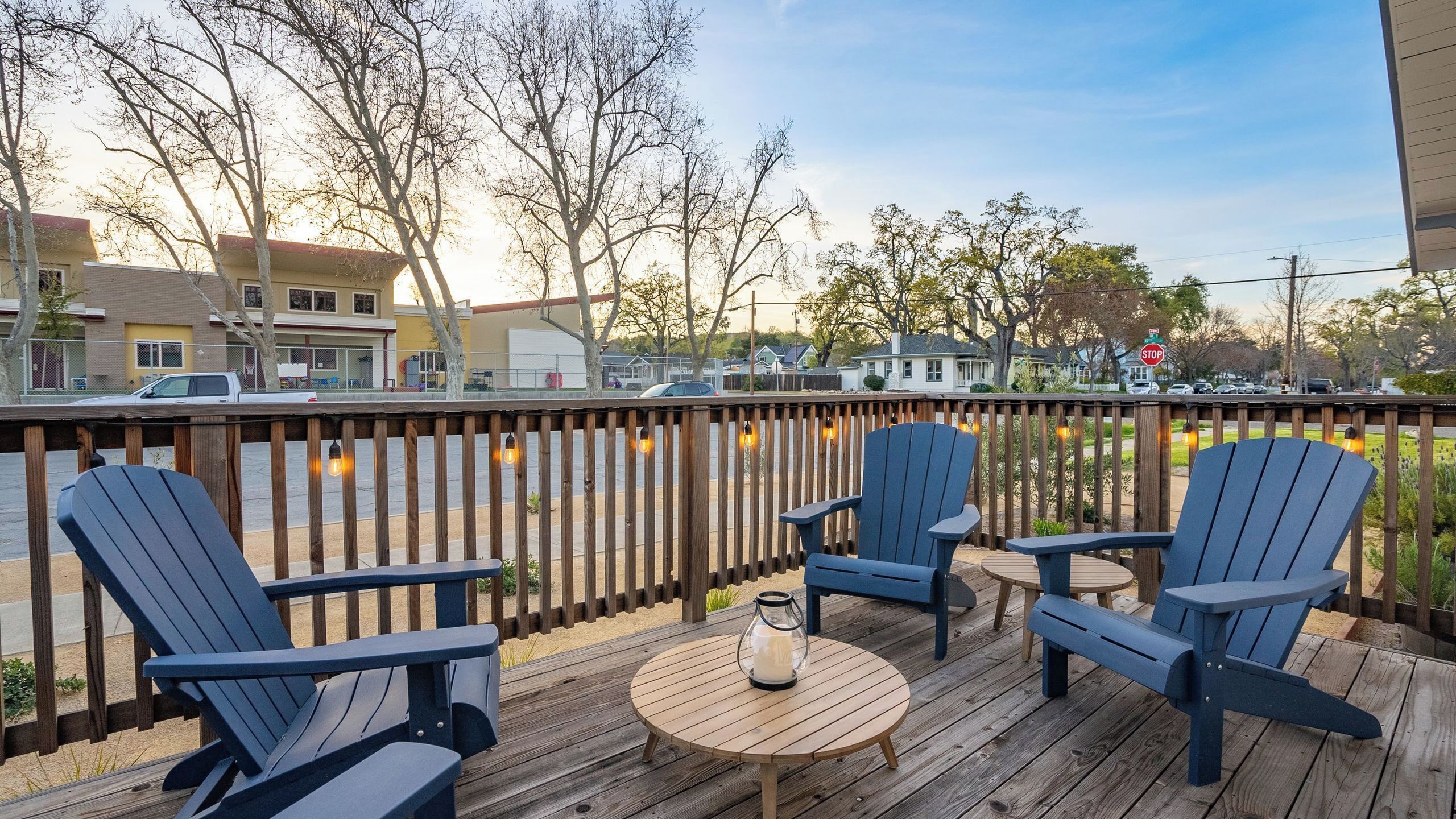 Terraza de madera con sillas Adirondack y una mesa redonda, con vista a una calle con edificios y coches estacionados.