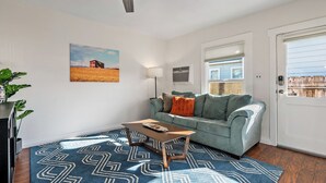 A living room with a blue patterned rug, a light blue sofa with cushions, a wooden coffee table, a painting of a field, and a door with a window. - Paso on Oak by Avantstay Cottage-style Home + Full Kitchen + Front Deck Near Downtown (Paso Robles)