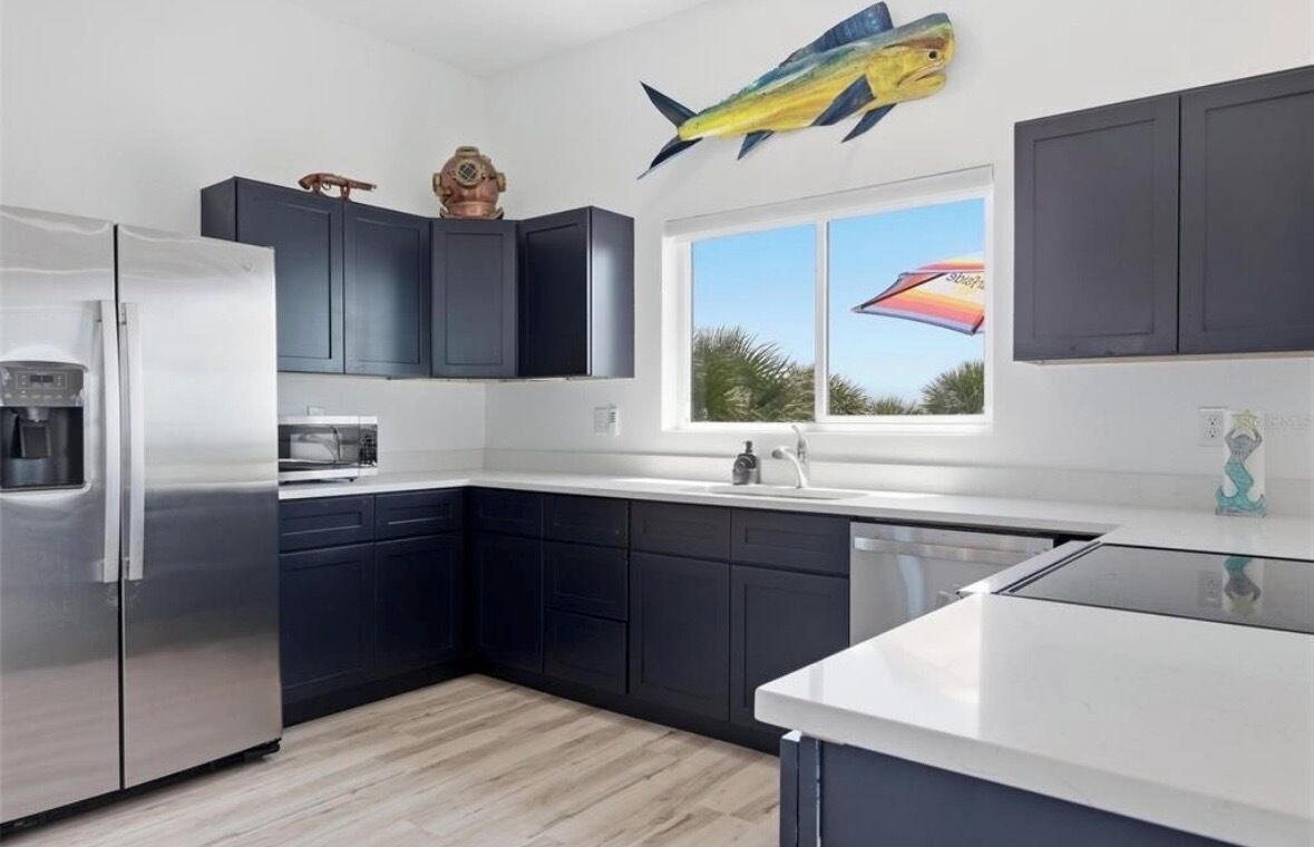 A modern kitchen with dark cabinets, a stainless steel refrigerator, and a window with a view of palm trees.