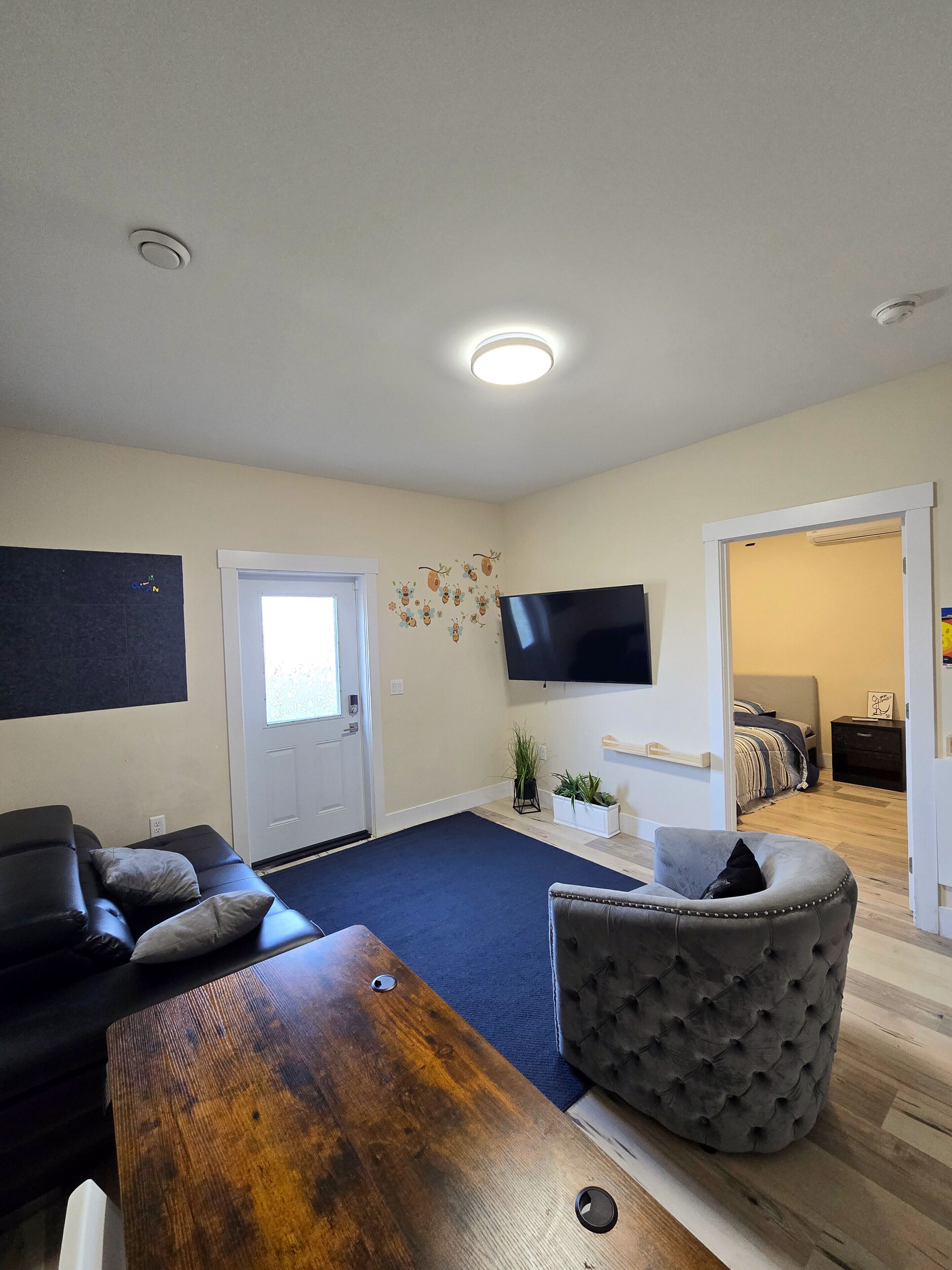 Un salón con un sofá de color oscuro, una mesa de centro de madera, un otoman tapizado de color gris, un televisor de pantalla plana montado en la pared y una puerta que da a otra habitación.