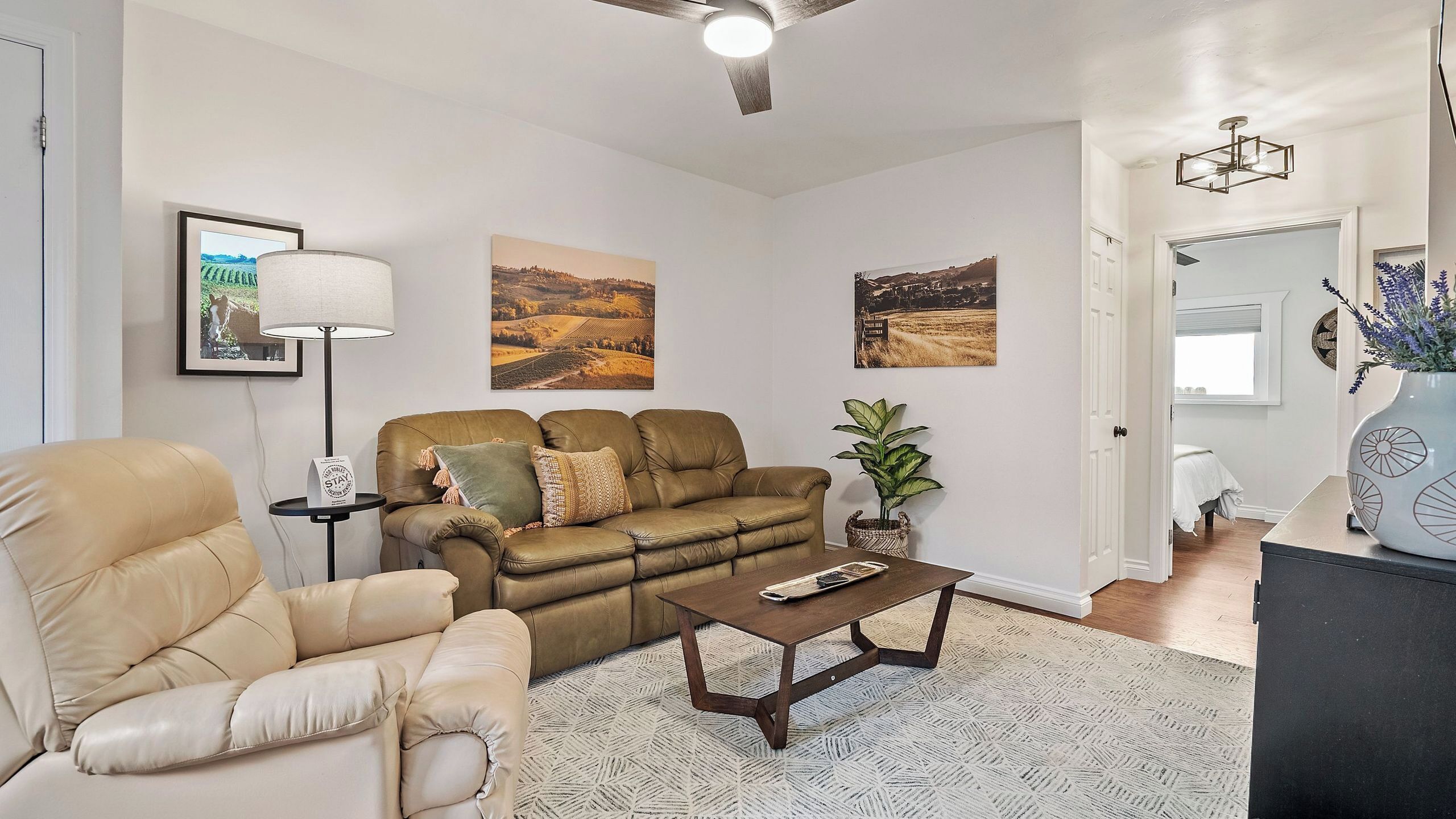 A living room with a sofa, armchair, coffee table, and a floor lamp. There are three framed pictures on the wall.