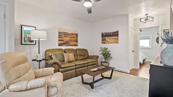 A living room with a sofa, armchair, coffee table, and a floor lamp. There are three framed pictures on the wall.