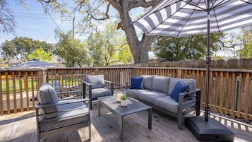 An outdoor patio with a wooden deck, a gray sofa with blue cushions, a metal table, and a striped umbrella.