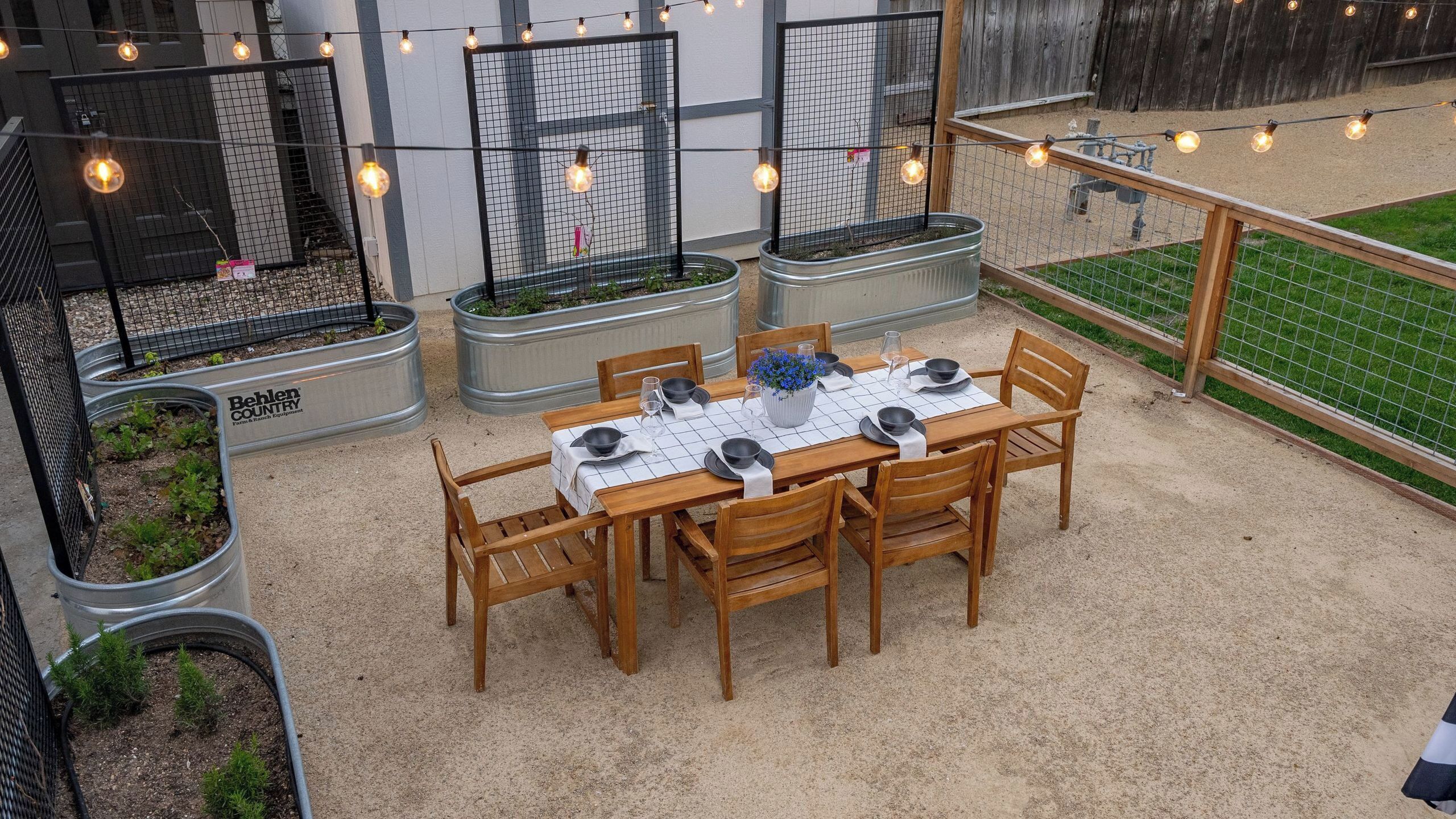 Un espace de restauration en plein air avec une table dressée pour quatre personnes, entourée de chaises, et une guirlande lumineuse suspendue au-dessus.