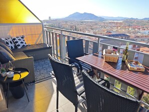 A balcony with wicker furniture, a table with a bottle and snacks, and a city view. - La Bella Vista 2-bedroom seaview AC apartment with elevator and garage (Puerto de Mazarrón)