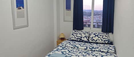 Un dormitorio con cama, dos almohadas, una mesita de noche con lámpara y una ventana con cortinas.