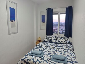 A bedroom with a bed, two pillows, a nightstand with a lamp, and a window with curtains. - La Bella Vista 2-bedroom seaview AC apartment with elevator and garage (Puerto de Mazarrón)