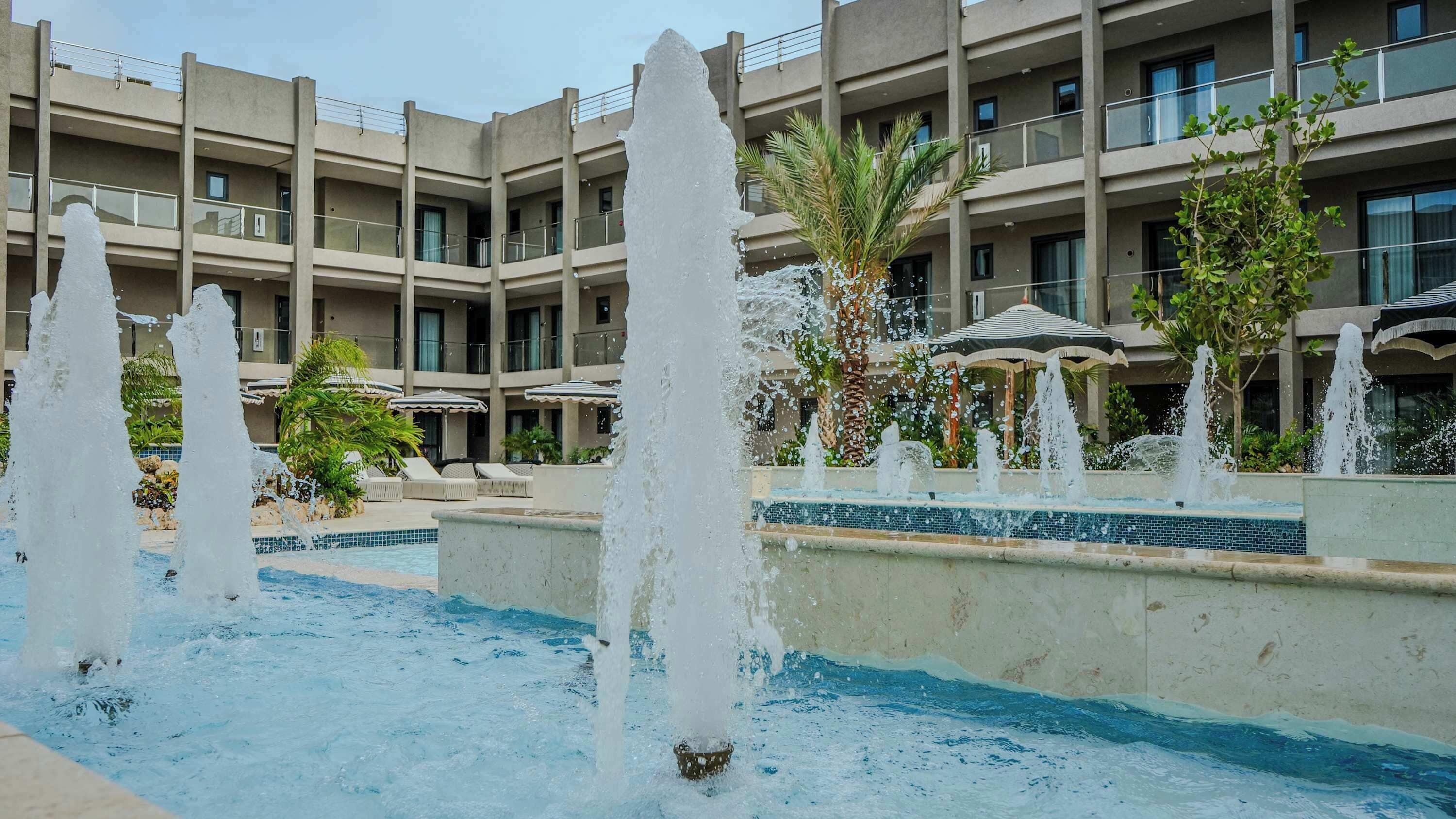 Une cour d’hôtel agrémentée de plusieurs fontaines, d’une piscine et d’un bâtiment doté de balcons.