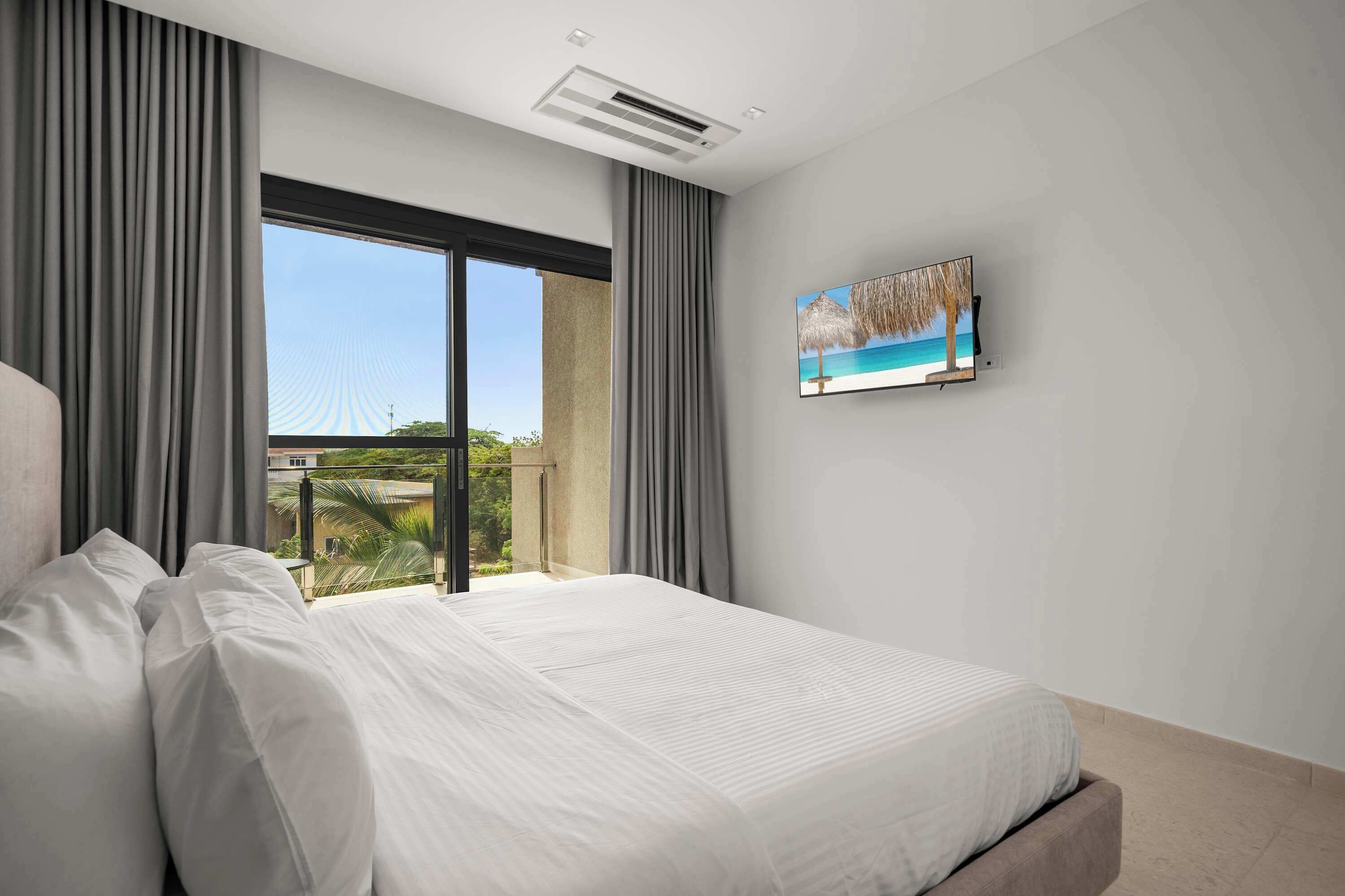 A hotel room with a large bed, a television mounted on the wall, and a balcony with a view of the outdoors.