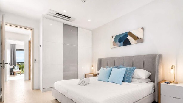 Un dormitorio moderno con una cama grande, mesitas de noche, una obra de arte en la pared y vistas al exterior a través de una puerta abierta.